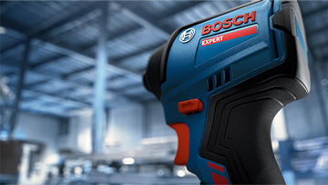 Modrý šroubovák Bosch s červeným tlačítkem, rozostřený v popředí, dílna v pozadí.