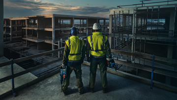 Zwei Bauarbeiter in Sicherheitskleidung stehen auf einer Baustelle mit unvollendeten Gebäuden.