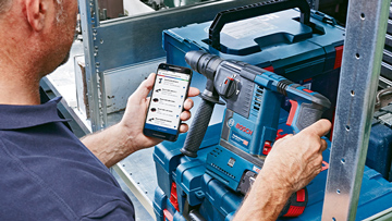 Řemeslník drží smartphone a dívá se na vrtačku na pracovním stole.
