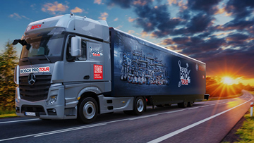 Šedý náklaďák jede po silnici, v pozadí je západ slunce s mraky.