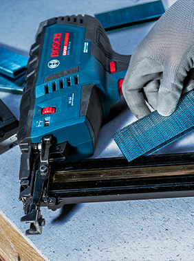 Osoba drží modrou vrtačku s červenou baterií v prostředí zpracování dřeva.