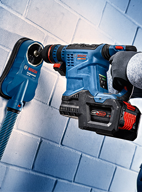 Modrý kladivový vrták vrtá do světlé zdi, s připojeným vysavačem.