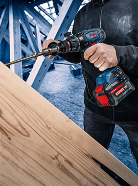 Řemeslník používá akumulátorovou vrtačku k opracování dřevěné desky. Pozadí s dřevěným rámem.