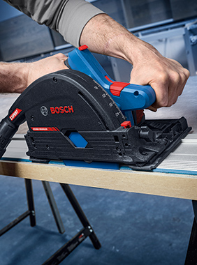 Ruka s modrou okružní pilou Bosch, která pracuje na dřevěné desce.