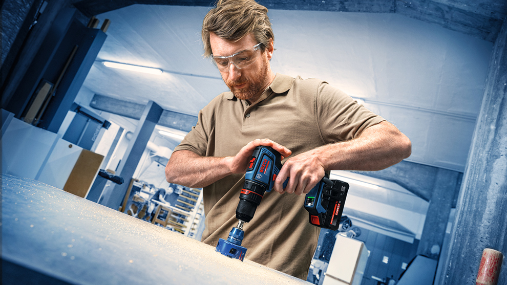 A man with glasses is drilling into a wooden board with a power drill in a workshop.