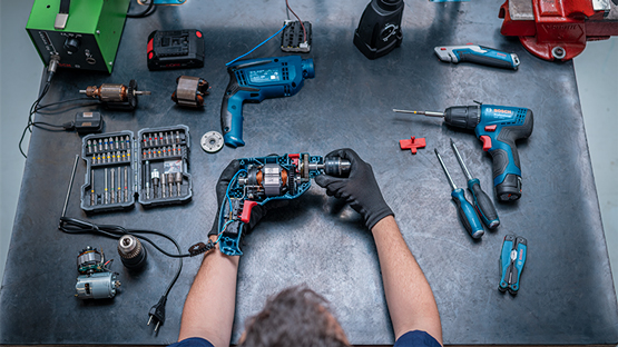 A worker wearing black gloves holds a disassembled Bosch Professional tool on a table. Various tools are scattered around him, including a blue cordless screwdriver, screwdrivers, and a box of bits. The environment is well-lit and tidy.