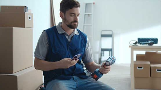 A bearded man is sitting on a box holding a blue Bosch Professional cordless drill in his hand. He is looking at his smartphone. In the background, moving boxes and a ladder are visible, and the scene appears bright and modern.