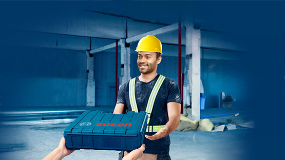 A man with a yellow helmet and reflective stripes holds a blue Bosch box in a construction site room.