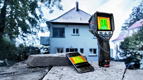 A black Bosch Professional infrared thermometer stands on a stone, next to a smartphone with thermal imaging.