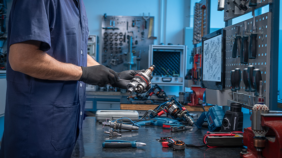 Um mecânico vestido com roupas de trabalho azuis segura uma ferramenta Bosch Professional na mão. Na mesa, há outras ferramentas, incluindo uma furadeira azul, chaves de fenda e uma caixa de ferramentas. Ao fundo, há ferramentas na parede e uma tela de computador mostrando desenhos técnicos. A iluminação é fria e azul, criando uma atmosfera profissional de oficina.