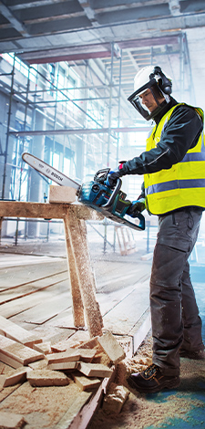 A worker in a yellow safety vest and helmet is using a chainsaw at a construction site. Wood chips are flying around.