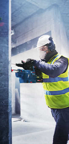 A worker in a yellow safety vest and helmet is using a blue Bosch Professional cordless nailer on a wall.