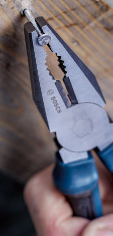 A gray Bosch Professional pliers grips a screw head, with a wooden surface in the background.