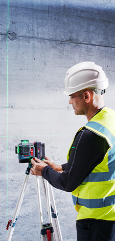 A man in a yellow safety vest and a white helmet is aligning a green laser measuring device on a tripod. The background is a gray concrete wall. The man stands sideways and focuses on aligning the device.