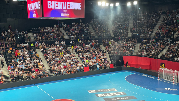 Salle pleine de fans, terrain de handball bleu et grand écran.