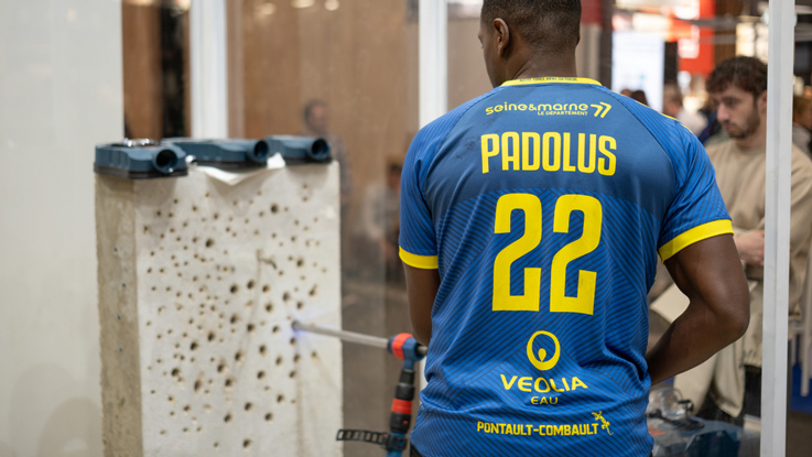 Un homme en maillot bleu se tient devant un mur perforé avec des outils.