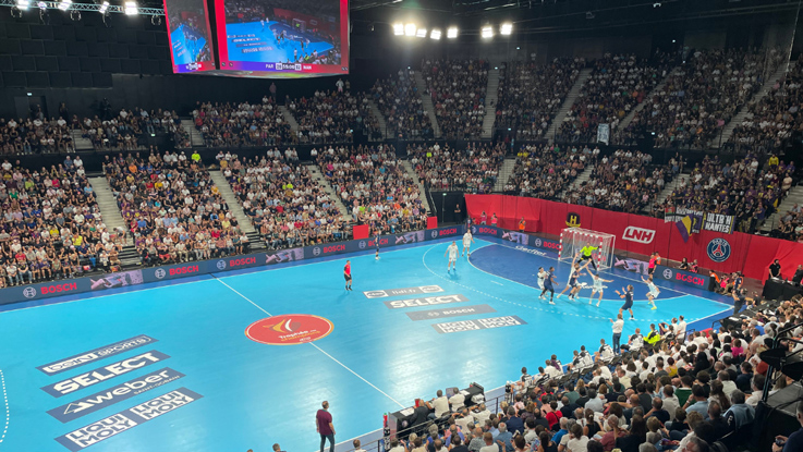Match de handball dans une salle avec un sol bleu, spectateurs, deux équipes en action.