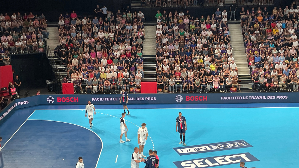 Match de handball sur un terrain bleu, tribune pleine de fans, publicité visible.
