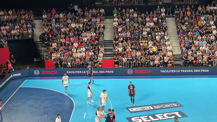 Match de handball dans une salle pleine avec un terrain bleu et des sponsors.