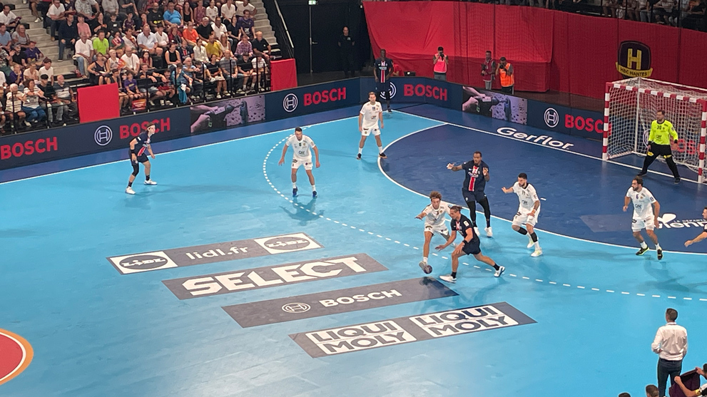Match de handball avec des joueurs en maillots bleus et blancs sur un terrain bleu.