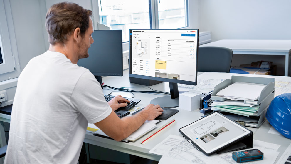 En man arbetar vid datorn i ett ljust kontor med byggplaner.