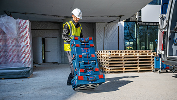 En arbejder i gul vest og hjelm løfter en blå værktøjskasse på byggepladsen. I baggrunden træpaller.