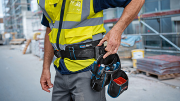 En arbejder i en gul sikkerhedsvest står på en byggeplads og rækker efter værktøjer i sin bælte-lomme.