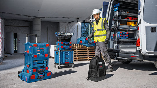 En arbejder i en sikkerhedsvest og hjelm står ved siden af en transporter, omgivet af blå Bosch Professional værktøjskasser.