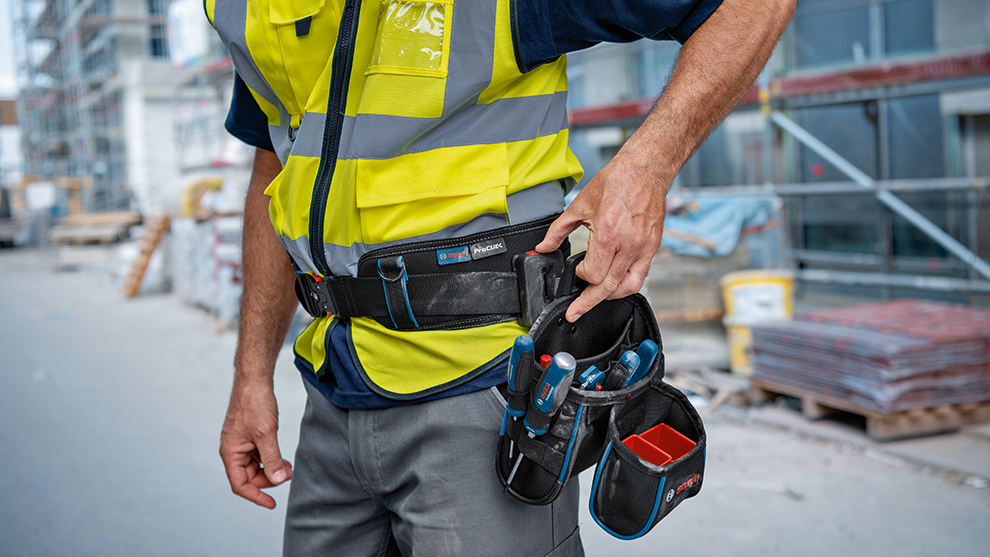 En håndværker i en gul advarselsvest griber ind i sin værktøjstaske for at tage værktøjer. I baggrunden er bygge materialer synlige.
