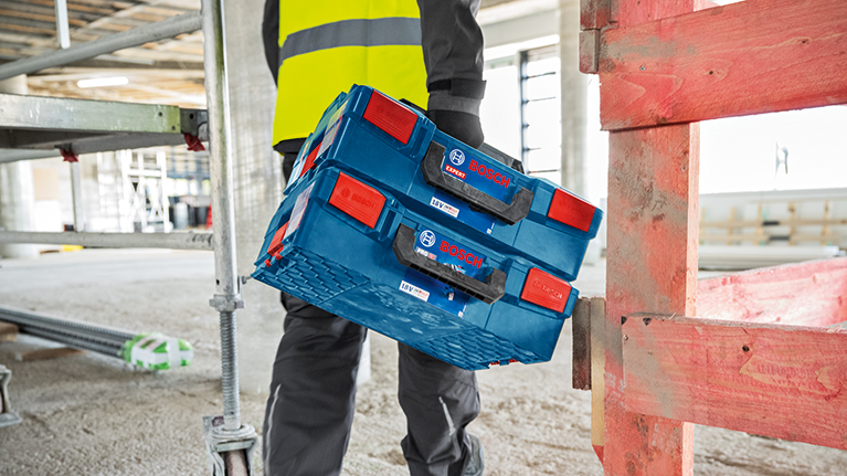 En arbejder i arbejdstøj løfter en blå Bosch Professional værktøjskasse med røde håndtag op på en anden. Omgivelserne er lyse og ser ud til at være en byggeplads.