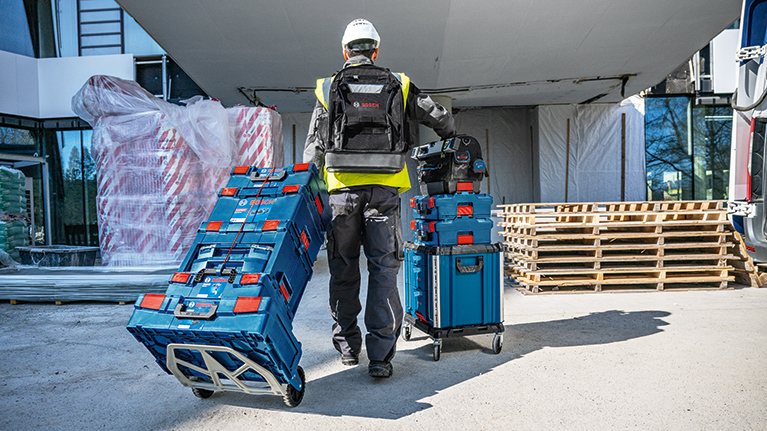 En arbejder i sikkerhedstøj skubber to blå værktøjskasser fra Bosch Professional på en byggeplads.