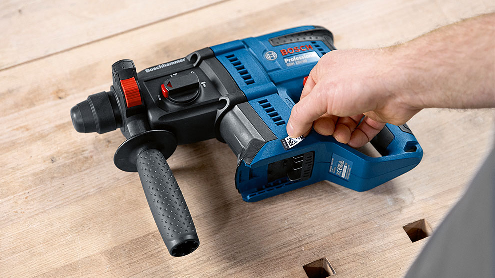 Un martello perforatore blu Bosch Professional è appoggiato su legno, una mano si allunga verso di esso.