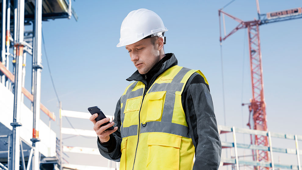 Ehitusworker kollases vestis ja kiivris vaatab oma nutitelefoni.