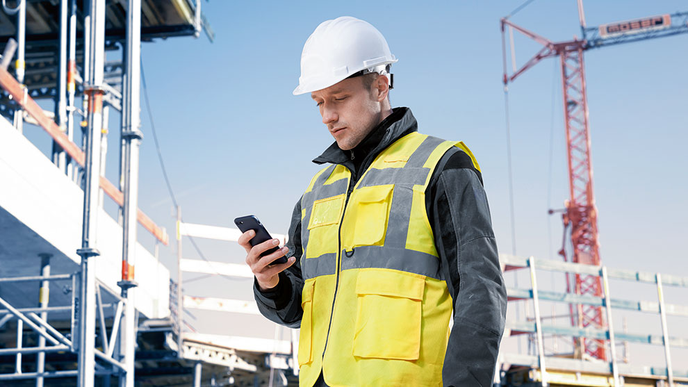 Ein Bauarbeiter in gelber Sicherheitsweste schaut auf sein Smartphone.