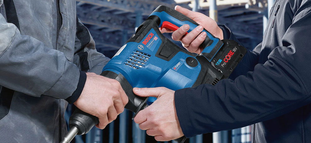 Två händer håller en blå Bosch Professional borrmaskin i en verkstad.