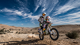 A motorcyclist in protective gear rides through a dry desert landscape.