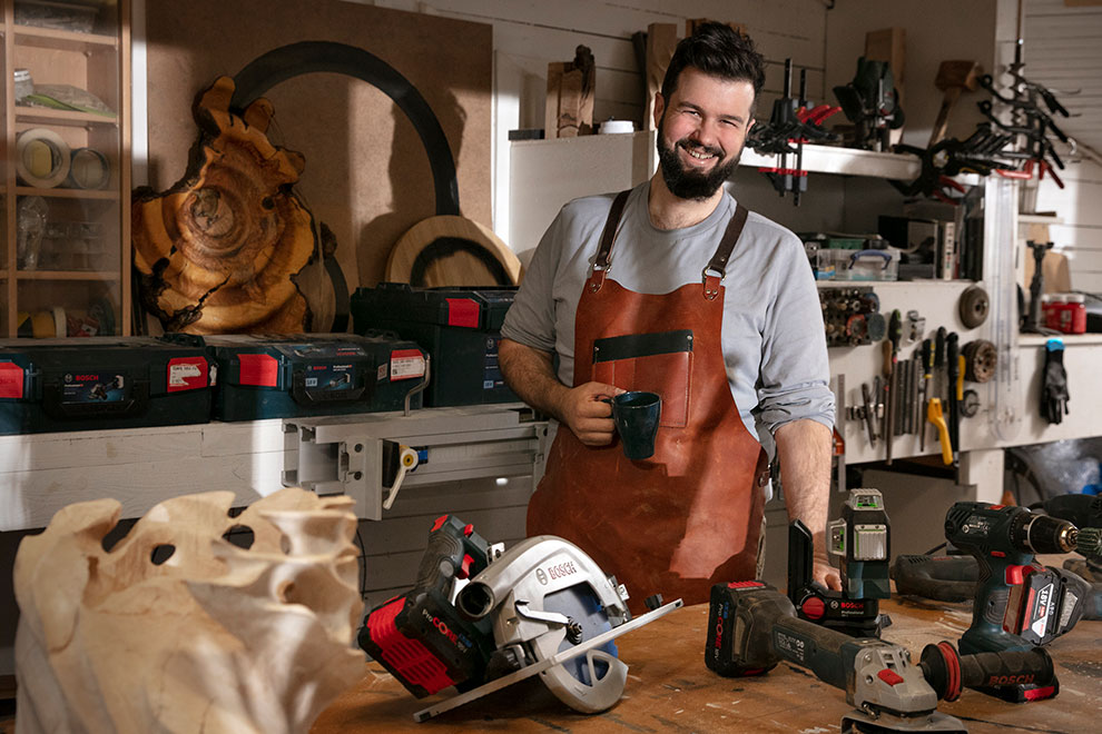 Mies ruskeassa esiliinissä seisoo pöydän ääressä Bosch-työkalujen kanssa.