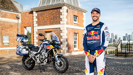 A man in a blue Red Bull racing suit stands next to a blue motorcycle in front of a historic building.