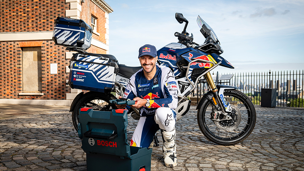 A smiling man in motorsport clothing sits in front of a blue Bosch box and a BMW motorcycle.