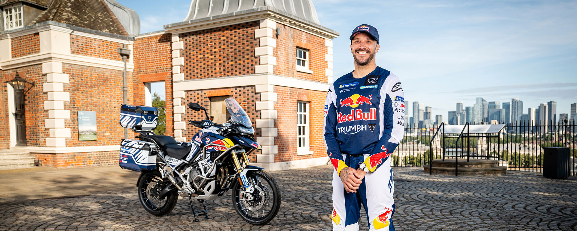 A man stands smiling in front of a Triumph motorcycle in Red Bull outfit, with a city view in the background.