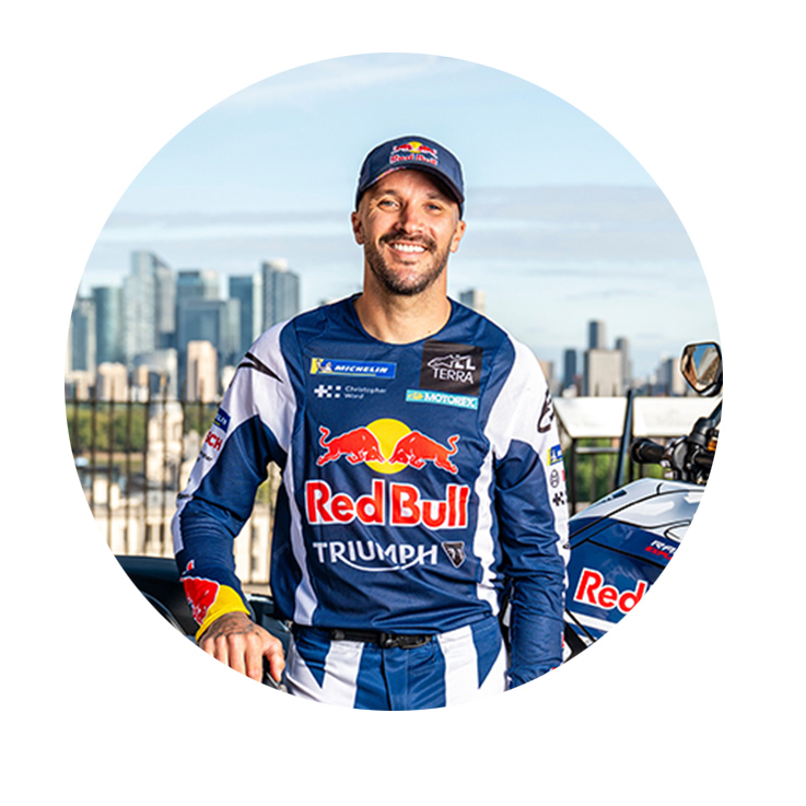 A smiling man in a blue Red Bull Racing outfit stands in front of a cityscape. Modern buildings are visible in the background.