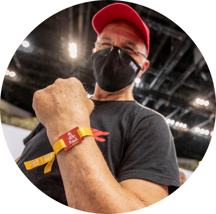 En man med mask visar sitt färgglada armband och ler.