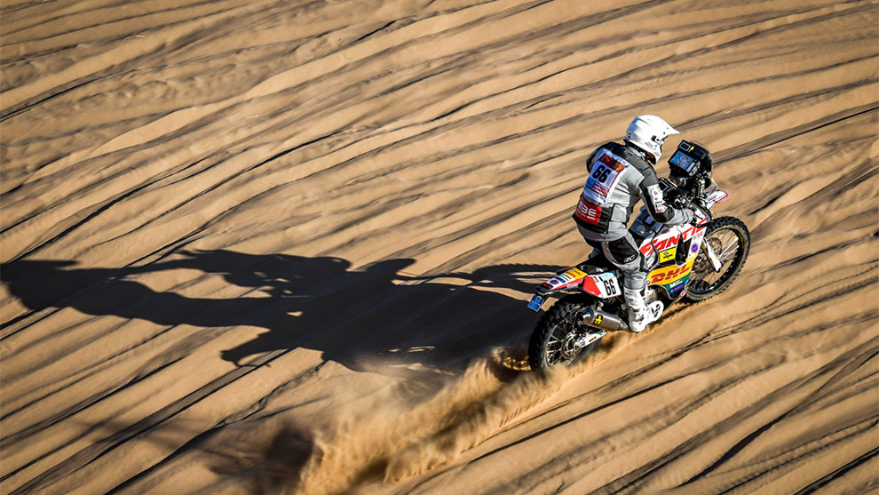 Motorcyklist på sand, lämnar spår, solen kastar långa skuggor.