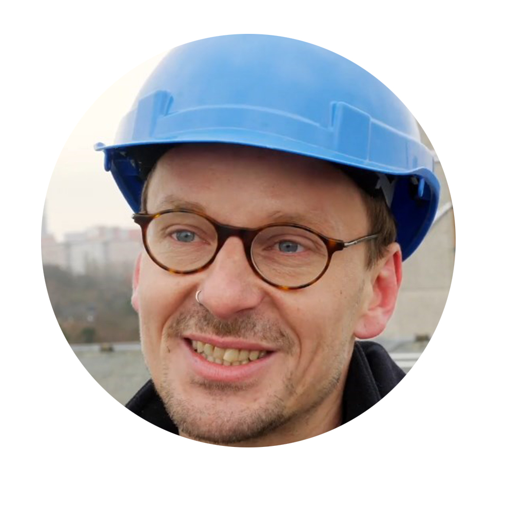 En man med blå säkerhetshjälm ler framför en suddig byggarbetsplatsbakgrund.