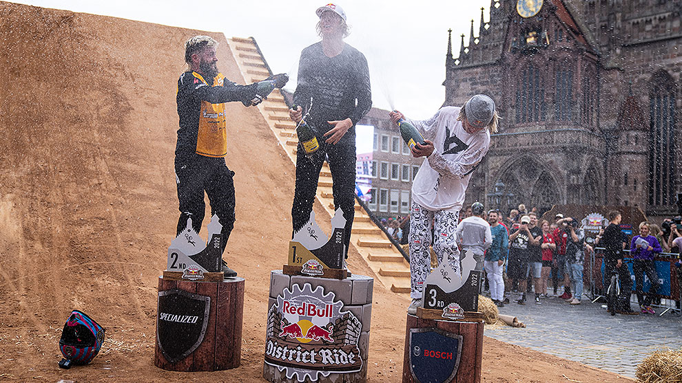 Three winners celebrate on podiums with champagne, a crowd in the background.