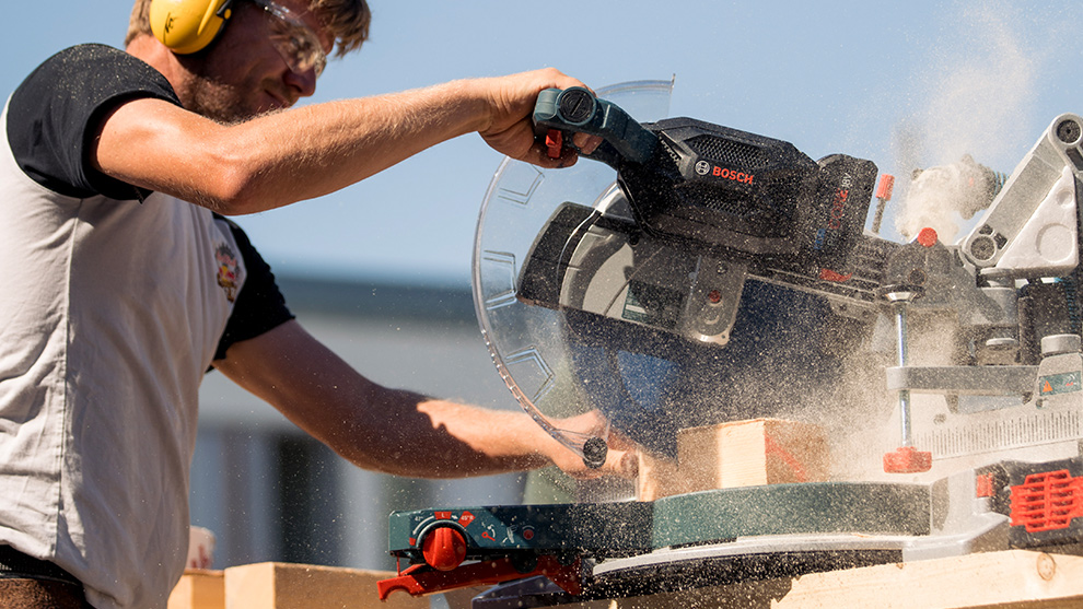 A man wearing hearing protection is cutting wood with a Bosch Professional miter saw.