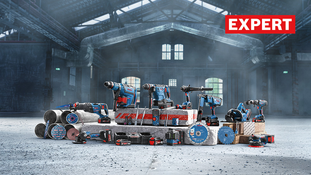 Een verzameling blauwe en zwarte elektrische gereedschappen op een grijze vloer in een industriehal. Te zien zijn boormachines, zagen en accu's, omringd door buizen en houten blokken. De omgeving is ruw en industrieel, met grote ramen en een gedempte, mistige sfeer.