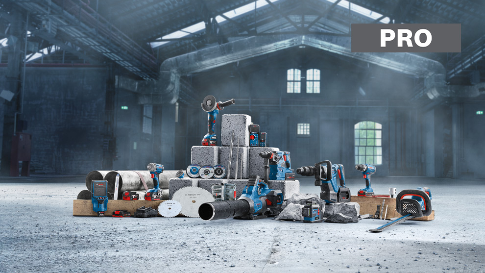 Een verzameling van elektrisch gereedschap en bouwmaterialen in een industriehal. Gereedschappen in blauw en rood, waaronder accuschroevendraaiers en zagen, zijn op grijze stenen en houten planken gerangschikt. De ruimte heeft hoge plafonds en grote ramen die diffuse verlichting bieden. De vloer is van beton en de sfeer is industrieel en ruw.