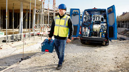 En arbetare i gul väst bär en blå verktygslåda på en byggarbetsplats.