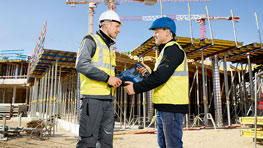 Två byggarbetare överlämnar ett verktyg till varandra på en byggarbetsplats.